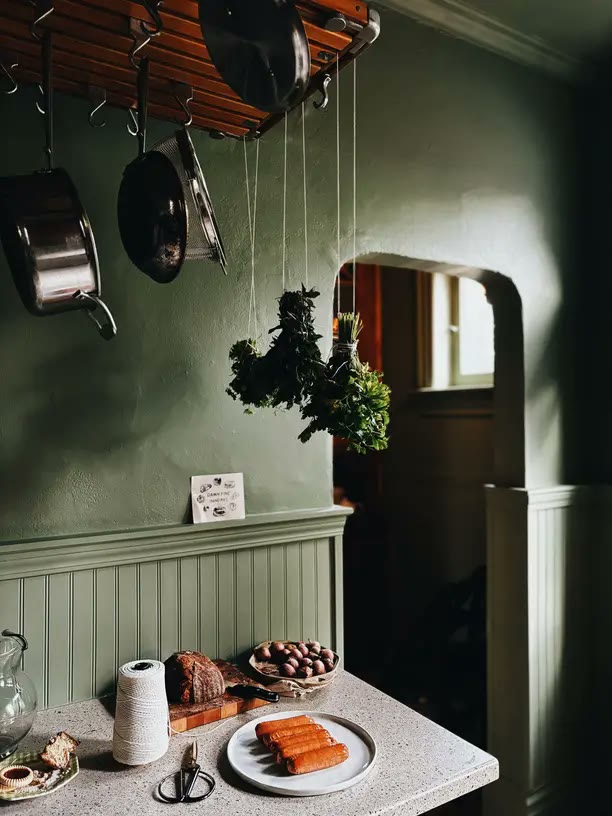 A dark green cottage kitchen with hanging copper pots and drying herbs