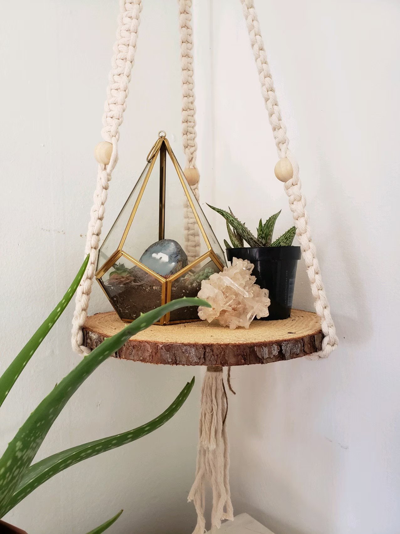 A cream macrame hanging shelf on a bark-edged wood slice, holding a gold terrarium, a crystal cluster and a succulent